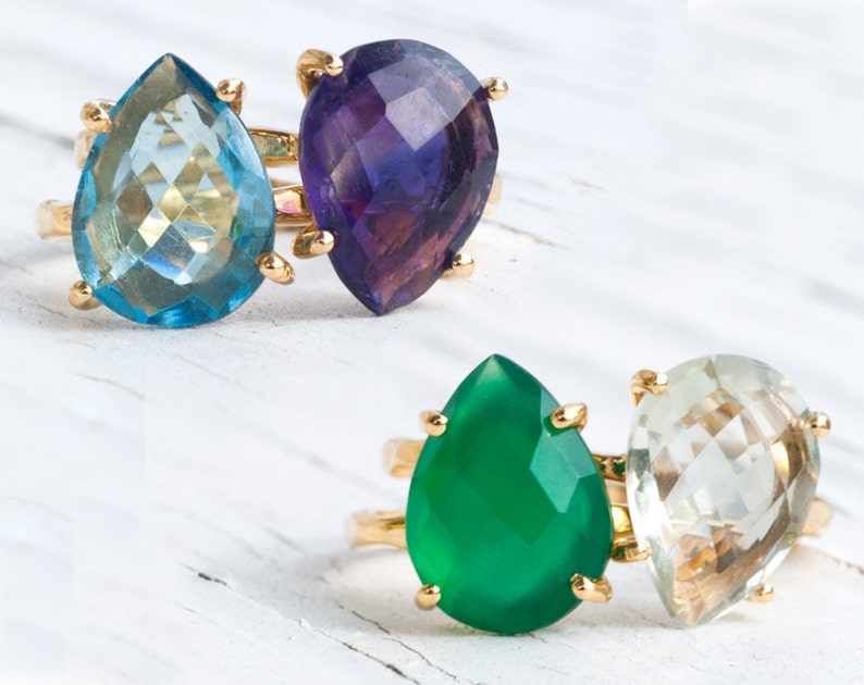 May include: Two gold rings with teardrop-shaped gemstones. The top ring features a light blue gemstone and a purple gemstone. The bottom ring features a green gemstone and a light green gemstone.