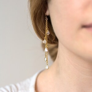 May include: Gold-toned dangle earrings featuring multiple chains and teardrop-shaped clear crystals. The earrings have a delicate design, with the crystals cascading down the chains. The jewelry is designed to be worn in the ear.