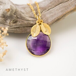 May include: A gold necklace with a purple amethyst gemstone pendant and two gold leaf charms. The necklace is on a wooden surface. AMETHYST is written below the image.
