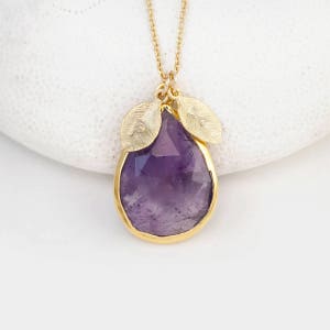 May include: A gold chain necklace with a teardrop-shaped amethyst pendant. The pendant is surrounded by two gold leaves with initials engraved on them.