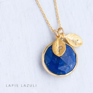 May include: Gold necklace with a blue lapis lazuli stone pendant and two gold leaf charms with the letters 'P' and 'A' engraved on them.  The pendant is round and has a faceted design.  The necklace is on a white background.  The text 'LAPIS LAZULI' is written below the necklace.