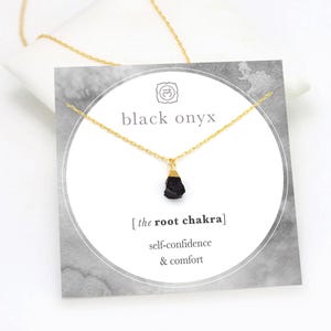 May include: A gold-colored chain necklace with a black onyx pendant. The pendant is a rough-cut stone with a gold-colored cap. The necklace is displayed on a card that reads "black onyx" and "the root chakra" with the words "self-confidence & comfort".