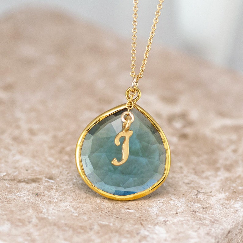 Pode incluir: Um colar de corrente de ouro com um pingente de pedra azul em forma de l&aacute;grima. O pingente tem uma borda folheada a ouro e um pequeno amuleto de ouro com a letra 'J' pendurado nele.