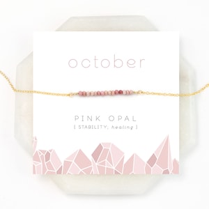 May include: October birthstone necklace with a pink opal stone bar pendant on a gold chain. The card reads "October" and "Pink Opal | Stability; healing".