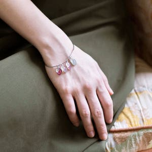 May include: A silver bangle bracelet with five charms. The charms are small, oval-shaped, and have a metallic finish. The charms are different colors, including pink, green, and silver.
