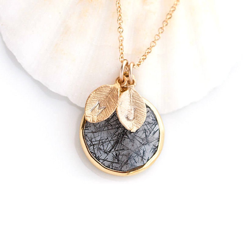 May include: Gold necklace with a round pendant featuring a black and gray stone with a gold rim. The pendant has two small gold leaves with the letters M and J engraved on them.
