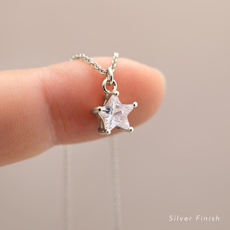 Puede incluir: Collar plateado con un colgante en forma de estrella. La estrella tiene una piedra central clara y facetada. La cadena es fina. El texto "Silver Finish" es visible en la parte inferior.