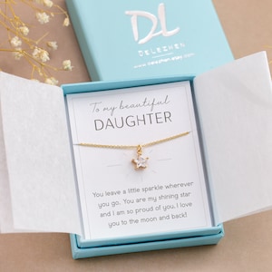 Puede incluir: Un collar dorado con un colgante en forma de estrella, presentado en una caja de regalo azul claro. La caja contiene una tarjeta con el texto "To my beautiful DAUGHTER" y un mensaje cari&ntilde;oso. La marca "DELEZHEN" es visible.