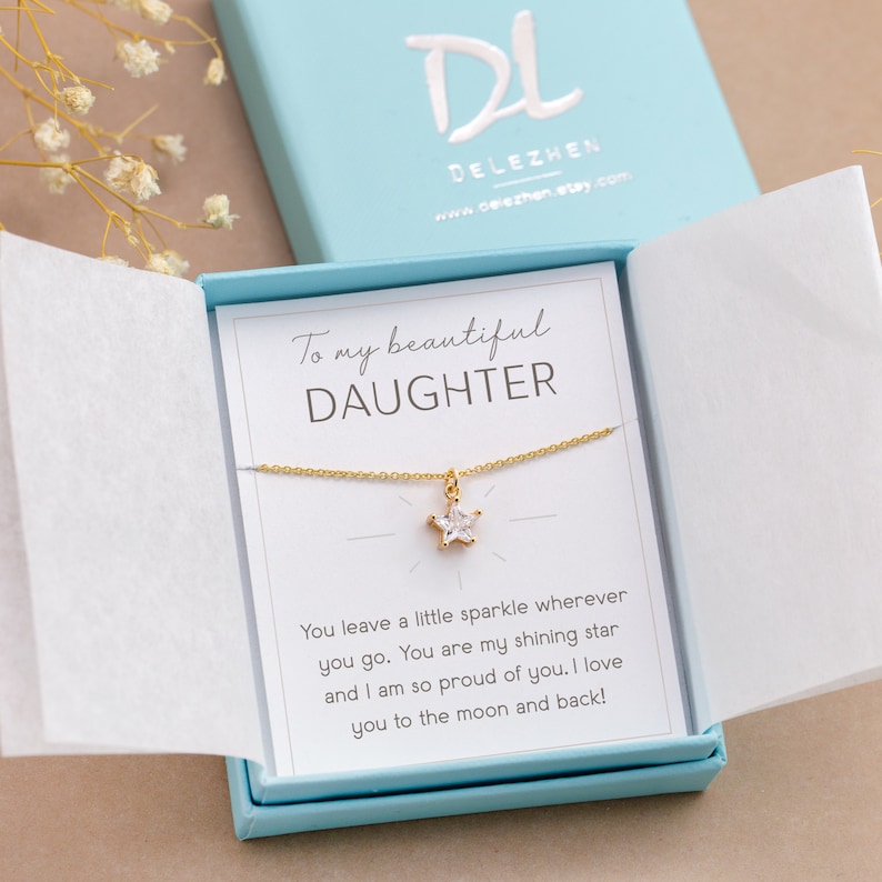 Puede incluir: Un collar dorado con un colgante en forma de estrella, presentado en una caja de regalo azul claro. La caja contiene una tarjeta con el texto "To my beautiful DAUGHTER" y un mensaje cari&ntilde;oso. La marca "DELEZHEN" es visible.