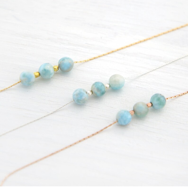 Pu&ograve; includere: Tre delicate collane con perline di pietre preziose blu e bianche su uno sfondo di legno bianco. Le collane sono realizzate con catene in oro, argento e oro rosa.
