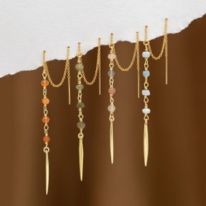K&ouml;nnte beinhalten: Vier Paar goldfarbene Ohrh&auml;nger. Jeder Ohrring hat eine Kette mit bunten Perlen und einem langen, spitzen Anh&auml;nger. Die Perlen sind in den Farbt&ouml;nen Orange, Gr&uuml;n, Rosa und Blau gehalten. Die Ohrringe h&auml;ngen vor einem wei&szlig;en, zerrissenen Papierhintergrund.