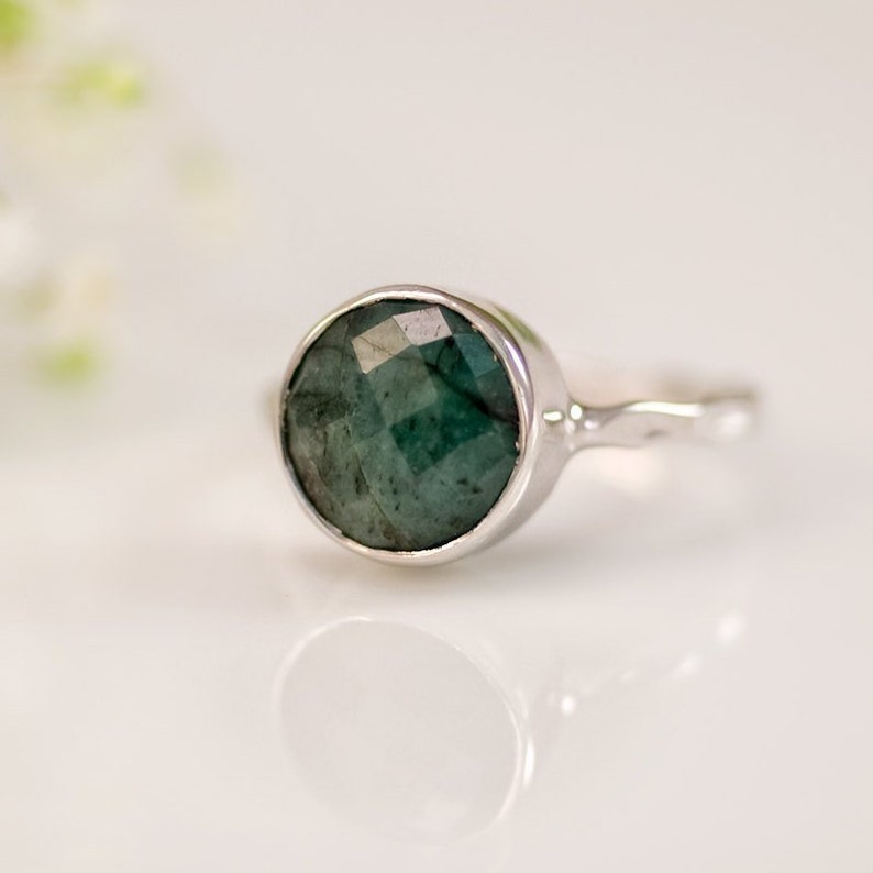 Pode incluir: Um anel de prata com uma gema de esmeralda verde. O anel tem uma banda simples e uma pedra redonda facetada.
