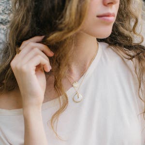 Pode incluir: Uma mulher vestindo uma camiseta branca com um colar de corrente dourada com um pequeno pingente redondo e um pingente maior em forma de l&aacute;grima com a letra 'S' nele.