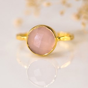 May include: A gold ring with a round, pale pink gemstone. The ring has a simple, delicate design.