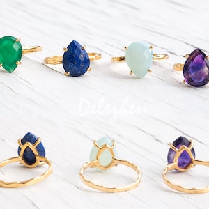 May include: Six gold rings with different colored gemstone settings. The gemstones are teardrop shaped and include green, blue, white, and purple.