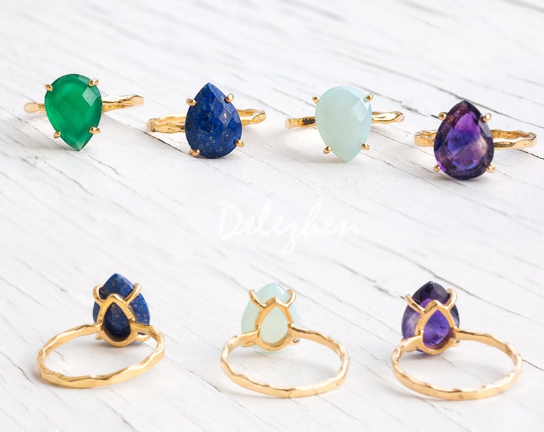 May include: Six gold rings with different colored gemstone settings. The gemstones are teardrop shaped and include green, blue, white, and purple.
