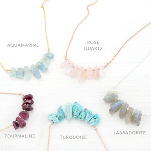 May include: Five necklaces with different colored gemstones. The necklaces are arranged in a row, with the gemstones forming a curved shape. The gemstones are aquamarine, rose quartz, turquoise, labradorite, and tourmaline.