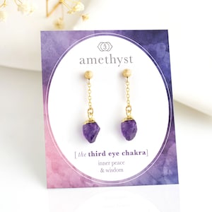 Op de afbeelding: Amethyst oorbellen met gouden kettingen en paarse kristallen druppels. De kaart vermeldt "de derde oog chakra" en "innerlijke vrede & wijsheid".