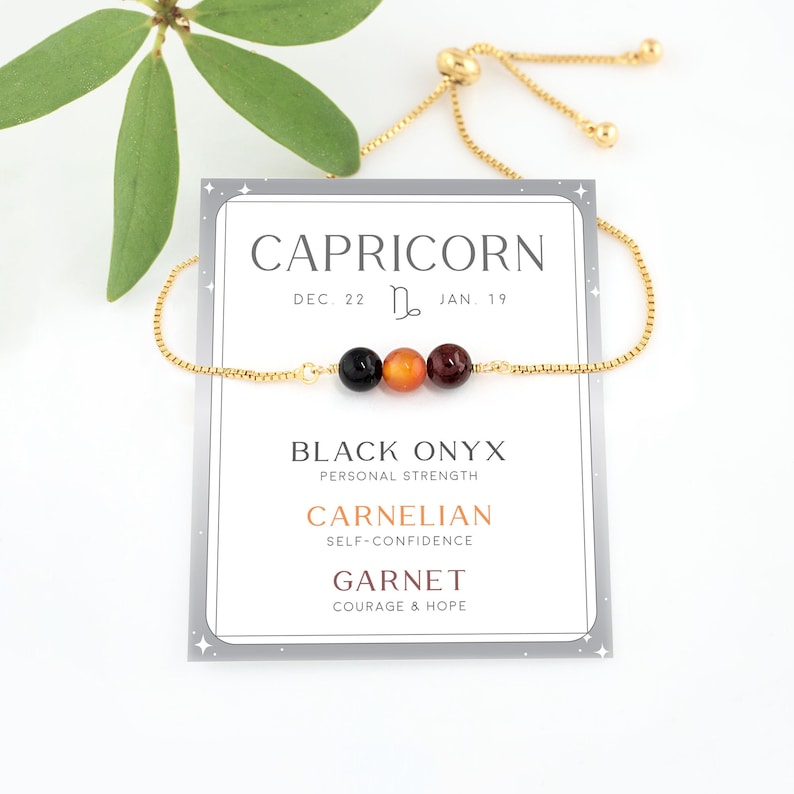 May include: A gold chain bracelet with three gemstone beads: black onyx, carnelian, and garnet. The card reads "Capricorn Dec. 22 - Jan. 19" and lists the stone meanings: Black Onyx - Personal Strength, Carnelian - Self-Confidence, Garnet - Courage & Hope.