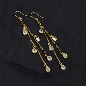 May include: Gold-toned dangle earrings featuring multiple chains with teardrop-shaped clear gemstones. The earrings have a hook closure and are displayed against a dark background.