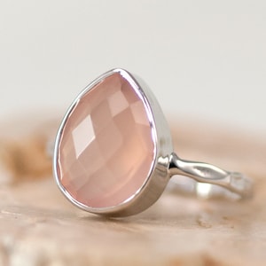 K&ouml;nnte beinhalten: Ein silberner Ring mit einem tropfenf&ouml;rmigen, facettierten, hellrosa Edelstein. Das Band des Rings hat ein strukturiertes Design. Der Edelstein ist in einer silbernen Fassung gefasst, was einen modernen Look erzeugt.