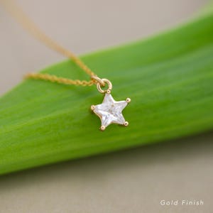 Puede incluir: Un collar dorado con un colgante en forma de estrella. El colgante es una piedra transparente y facetada engastada en metal dorado. El collar se muestra sobre un fondo verde. Las palabras "Gold Finish" son visibles en la esquina inferior derecha.