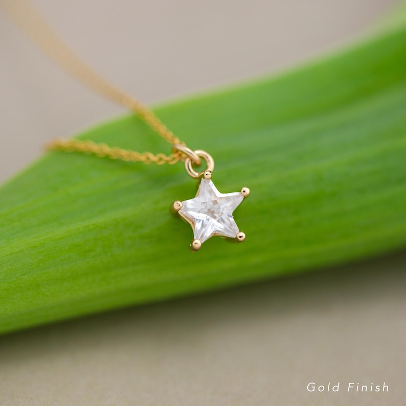 Puede incluir: Un collar dorado con un colgante en forma de estrella. El colgante es una piedra transparente y facetada engastada en metal dorado. El collar se muestra sobre un fondo verde. Las palabras "Gold Finish" son visibles en la esquina inferior derecha.