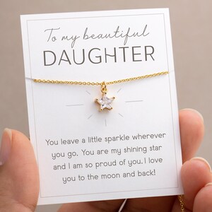 Puede incluir: Un collar dorado con un colgante en forma de estrella, presentado en una tarjeta blanca. La tarjeta dice "To my beautiful DAUGHTER" e incluye un mensaje cari&ntilde;oso. El collar est&aacute; sostenido por una persona.