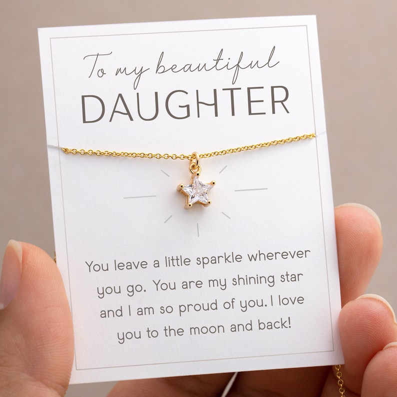 Puede incluir: Un collar dorado con un colgante en forma de estrella, presentado en una tarjeta blanca. La tarjeta dice "To my beautiful DAUGHTER" e incluye un mensaje cari&ntilde;oso. El collar est&aacute; sostenido por una persona.