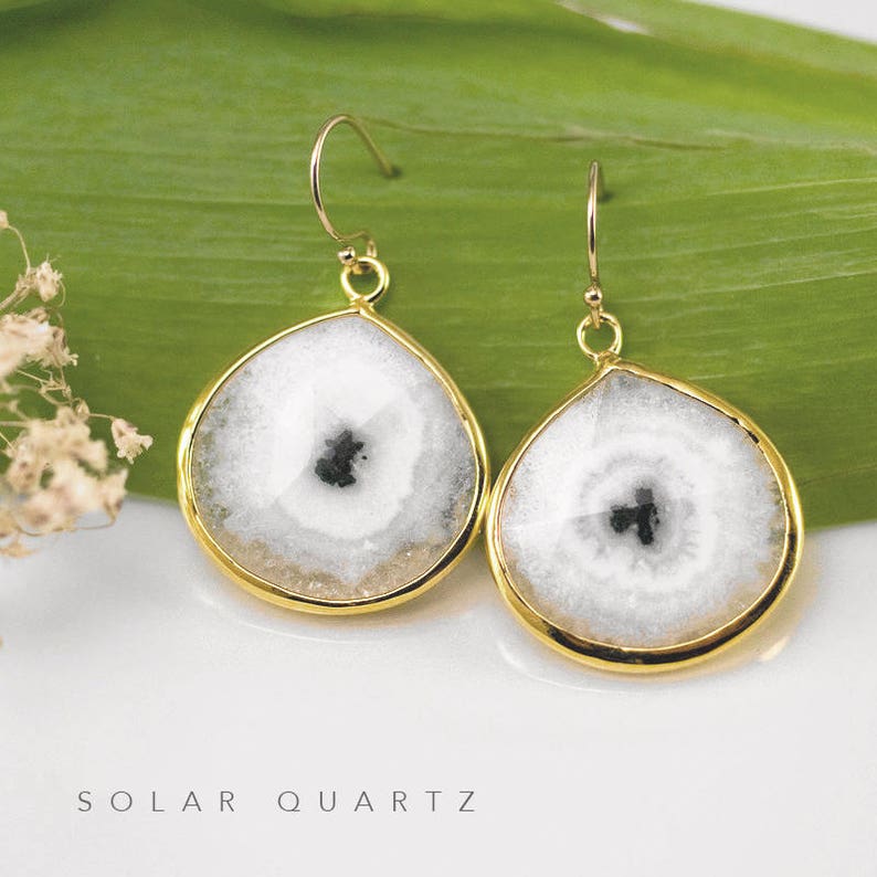 May include: A pair of gold earrings with white and black solar quartz stones. The stones are round and have a teardrop shape. The earrings are hanging from gold hooks.