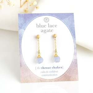 Op de afbeelding: Een paar gouden oorhangers met blauwe agaatstenen. De oorbellen bevinden zich op een kaart met de tekst "blauwe agaat" en "[de keelchakra] kalme en zelfverzekerde communicatie".