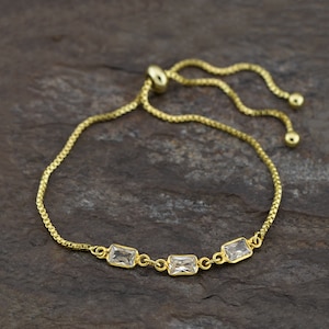 Puede incluir: Una pulsera dorada con tres piedras preciosas rectangulares y transparentes. La pulsera tiene una cadena delicada y un cierre deslizante ajustable. Las piedras preciosas están engastadas en un metal dorado. La pulsera se muestra sobre una superficie oscura y texturizada.