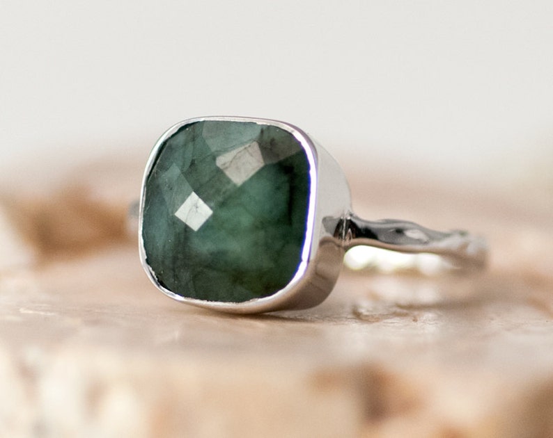 Pode incluir: Um anel de prata com uma pedra preciosa verde quadrada. O anel tem uma banda texturizada.