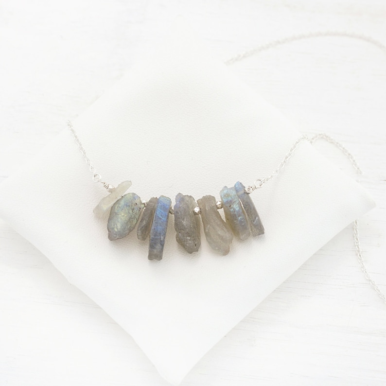 May include: A silver chain necklace with a cluster of raw labradorite stones hanging from it. The stones are a mix of gray and blue with iridescent flashes.