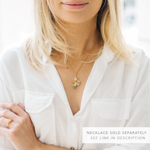 May include: A woman wearing a white button-down shirt with a gold necklace featuring a cluster of oval-shaped stones in shades of pink, blue, and gray. The text "NECKLACE SOLD SEPARATELY SEE LINK IN DESCRIPTION" is at the bottom of the image.