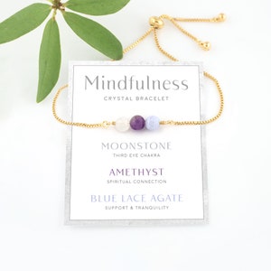 May include: A gold chain bracelet with three beads: a white moonstone, a purple amethyst, and a blue lace agate. The bracelet is adjustable and has a card with the text "Mindfulness Crystal Bracelet" and the names and properties of each stone.
