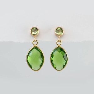 May include: A pair of gold earrings with two green gemstones each. The top stone is round, and the bottom stone is an elongated oval shape. The earrings have a simple, elegant design, suitable for everyday wear.