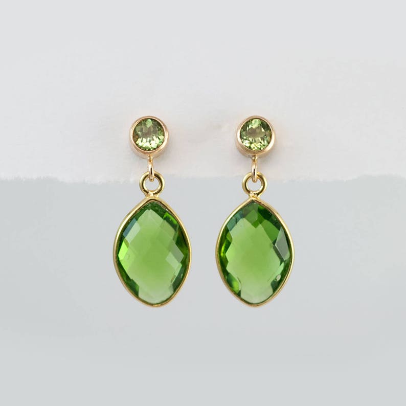 May include: A pair of gold earrings with two green gemstones each. The top stone is round, and the bottom stone is an elongated oval shape. The earrings have a simple, elegant design, suitable for everyday wear.