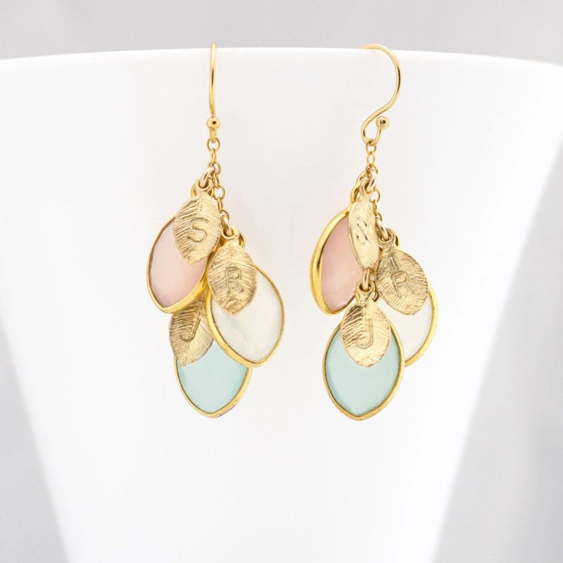 May include: Gold dangle earrings with multiple teardrop-shaped stones in pink, white, and light blue. Each stone has a gold leaf charm with a letter engraved on it.
