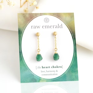 Op de afbeelding: Ruwe smaragd hangende oorbellen met gouden kettingen en een groene steen. De kaart heeft de tekst "raw emerald" en "[the heart chakra] love, harmony & communication".