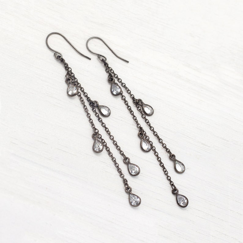 May include: A pair of long, dangling earrings with a dark gray finish. Each earring features multiple chains with teardrop-shaped, clear gemstones. The earrings are displayed on a white surface.
