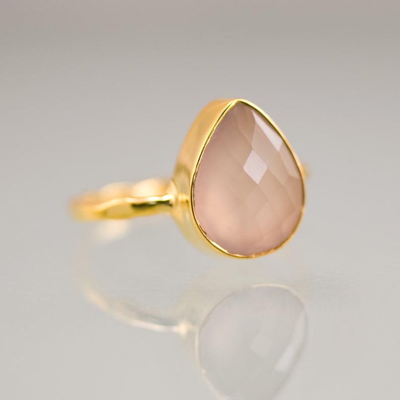 K&ouml;nnte beinhalten: Ein goldener Ring mit einem tropfenf&ouml;rmigen, facettierten, hellrosa Edelstein. Der Ring hat ein schlichtes, elegantes Design mit einem glatten, polierten Goldband. Der Edelstein ist in einer goldenen L&uuml;nette gefasst.