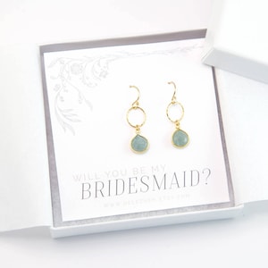 May include: A white card with the text "Will you be my bridesmaid?" and a website address. Two gold earrings with light blue teardrop-shaped stones are displayed on the card.