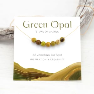 Puede incluir: Una tarjeta con el texto "Green Opal, Stone of Change, Comforting Support, Inspiration & Creativity" y una cadena dorada con cuentas de &oacute;palo verde.
