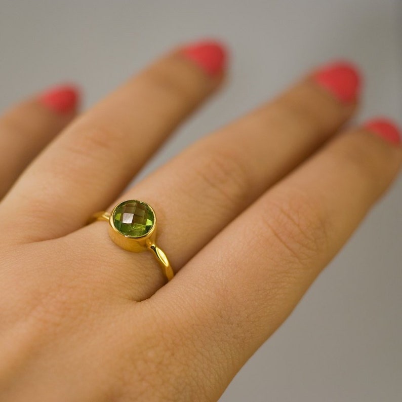 Pu&ograve; includere: Un anello d'oro con una gemma verde chiaro, rotonda e sfaccettata. L'anello ha una semplice montatura a castone ed &egrave; mostrato su un dito. Lo sfondo &egrave; di un colore neutro e morbido.