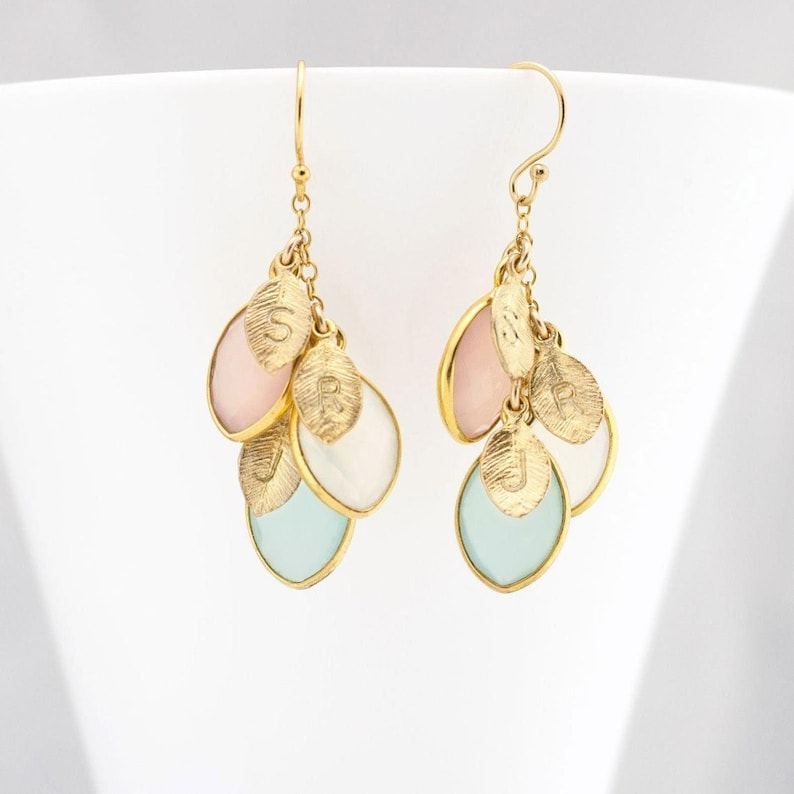 May include: Gold dangle earrings with multiple teardrop-shaped charms. The charms are in shades of pink, white, and blue, and each has a gold leaf with a letter engraved on it.