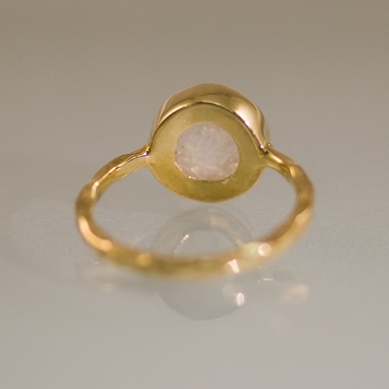 Puede incluir: Un anillo de oro con una piedra ovalada blanca engastada en un engaste de bisel. La banda del anillo es texturizada y tiene un acabado martillado.