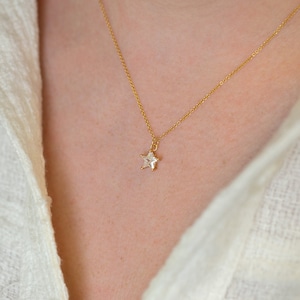 Puede incluir: Un collar dorado con un colgante en forma de estrella. El colgante tiene una gema transparente de corte cuadrado engastada en un marco dorado. El collar se muestra sobre un fondo blanco.