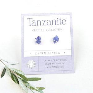 Puede incluir: Pendientes de cristal de Tanzanita en un engaste de oro. Los pendientes están en una tarjeta blanca con el texto "Tanzanite Crystal Collection" y "Crown Chakra". La tarjeta también incluye el texto "Chakra of Intuition. Sense of Purpose. And Connection."