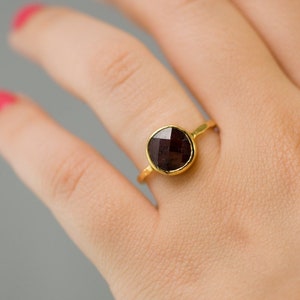 K&ouml;nnte beinhalten: Ein goldener Ring mit einem dunkelroten Edelstein. Der Ring befindet sich an einem Finger.